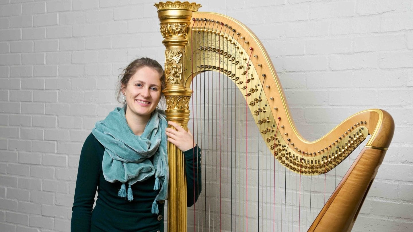 A woman in a blue scarf stands next to a harp.