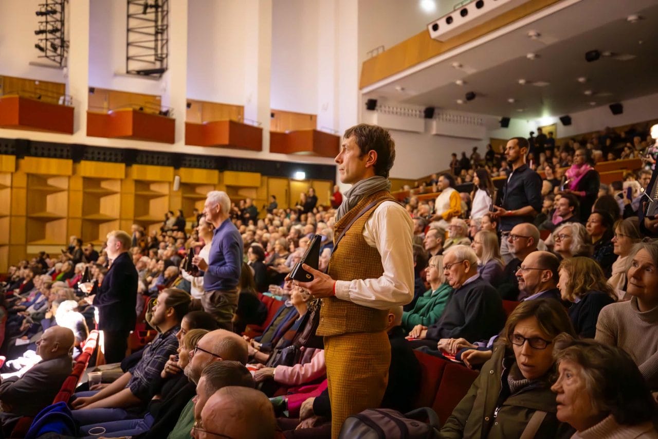 Audience members holding metronomes stand in busy concert hall.