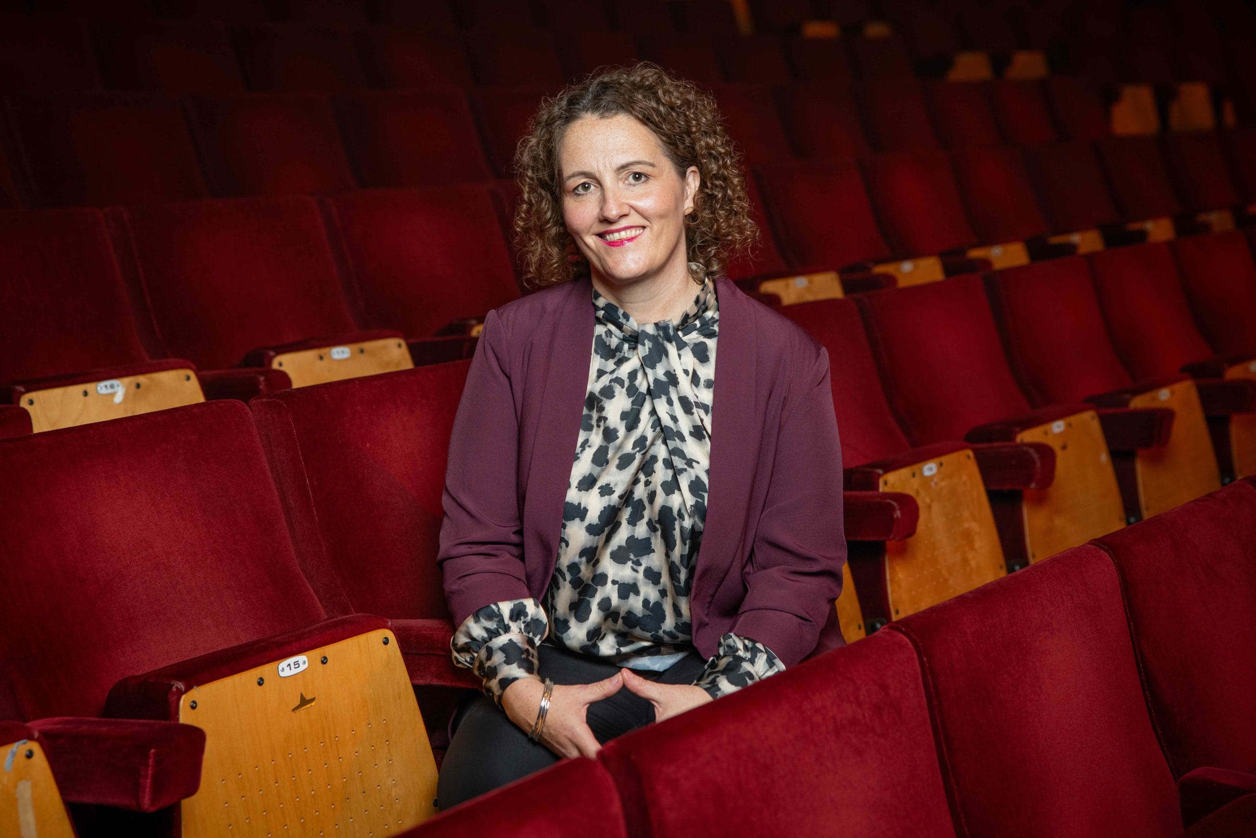 A smiling woman photographed in the red seats of a concert hall.
