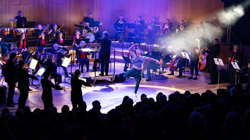 A dancer is captured mid-leap on stage, with musicians arranged around him.