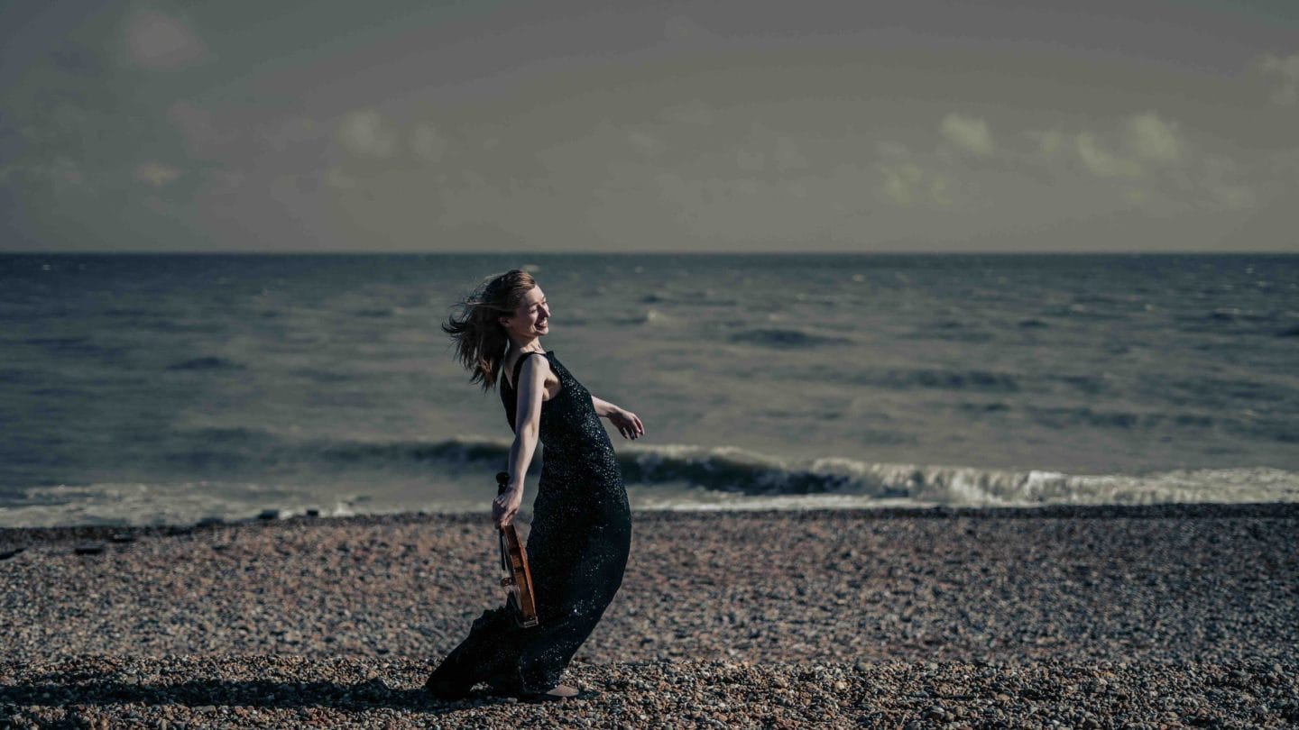 Woman with a violin in one hand leans backwards on a pebble beach.