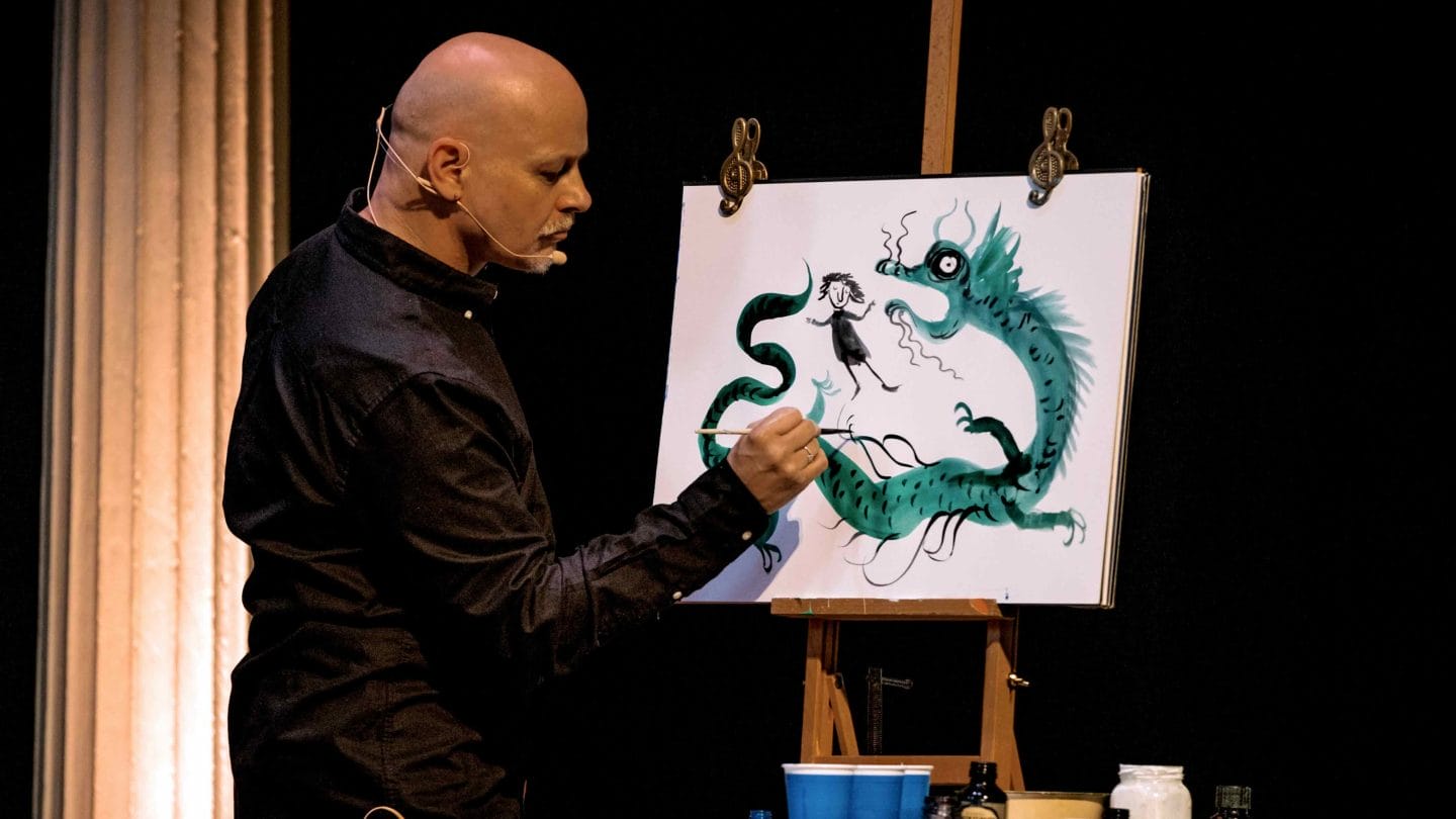 A man with a headset microphone paints a picture of a dragon on an easel.
