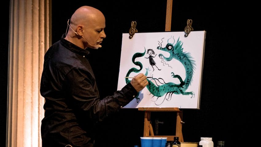 A man with a headset microphone paints a picture of a dragon on an easel.