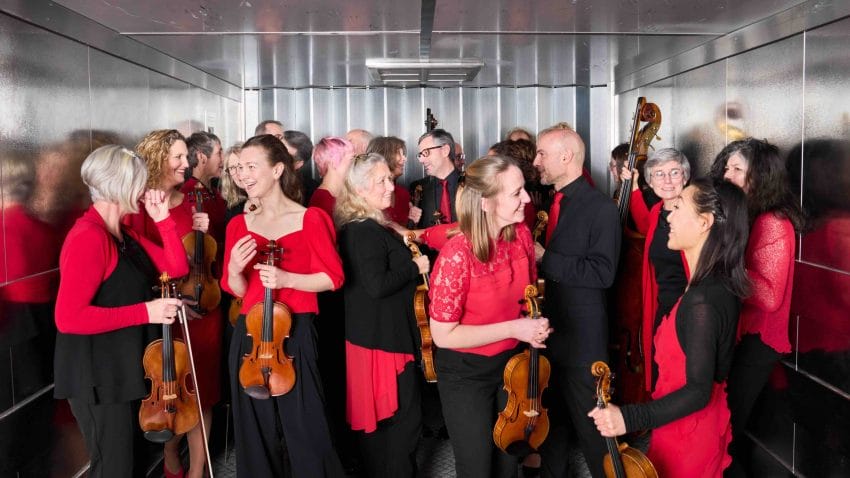 Musicians in black and red gather inside a large lift with their instruments.