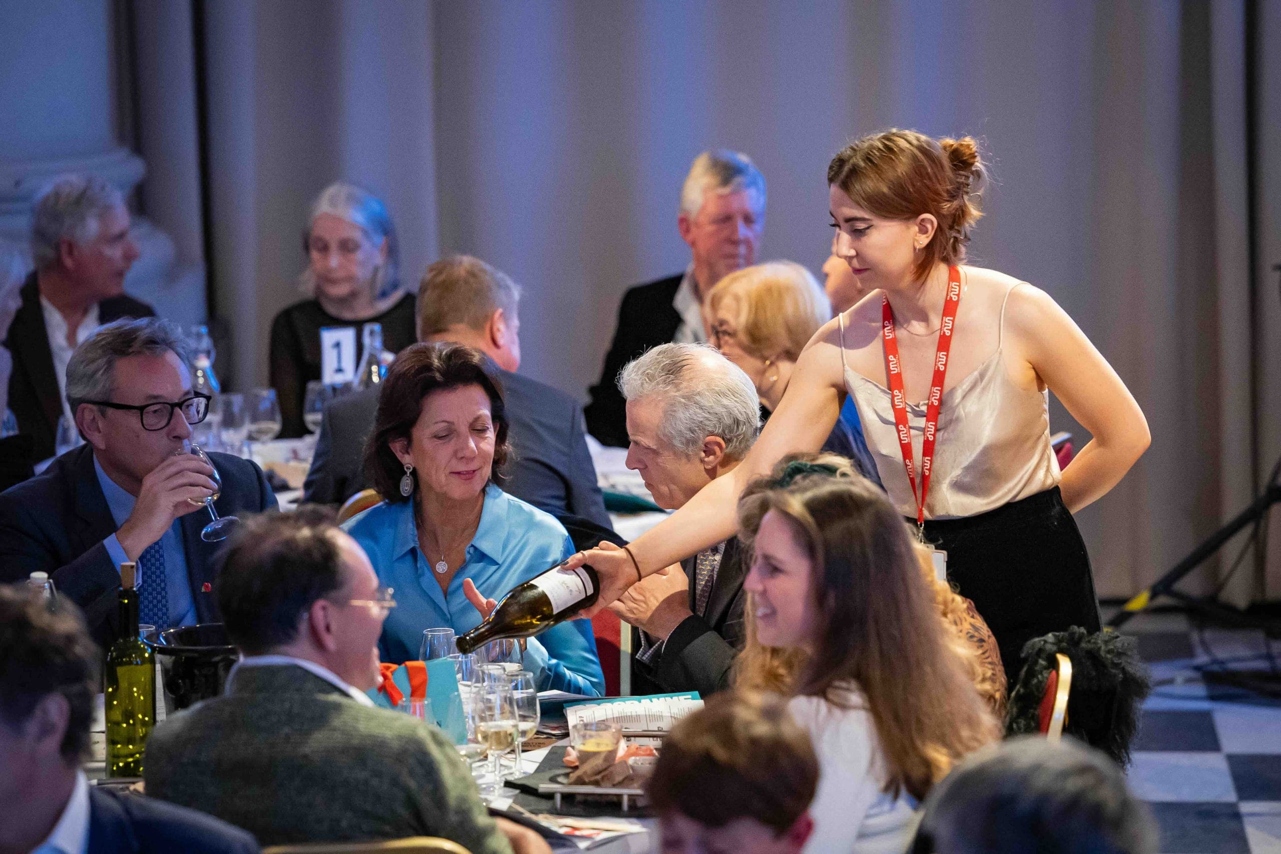 Woman in lanyard serves wine to seated guests.