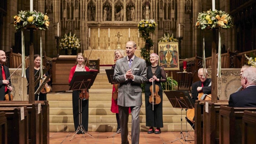 Prince Edward speaks with musicians stnading behind him with their instruments.