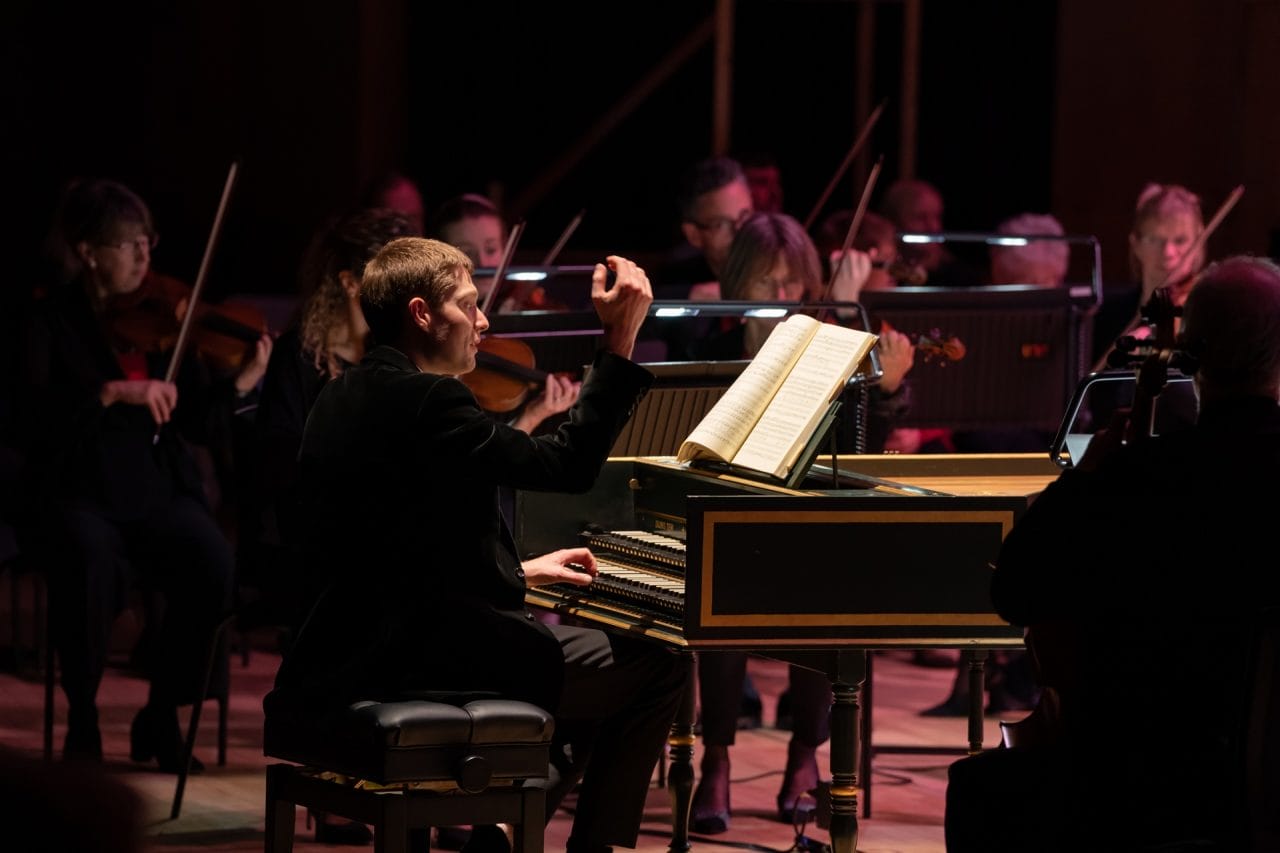 A man raises his hand to conduct an orchestra, sitting at a harpsichord.