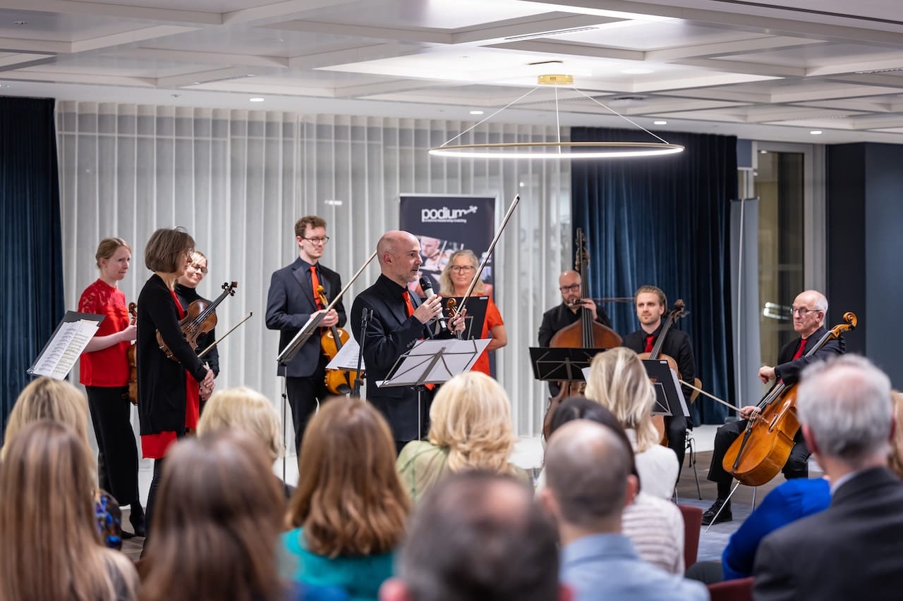 Man holding a violin addresses and audience with a microphone.