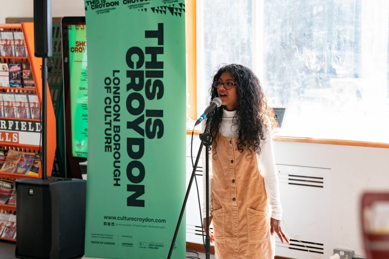 A young girl in a pinafore sings into a microphone.