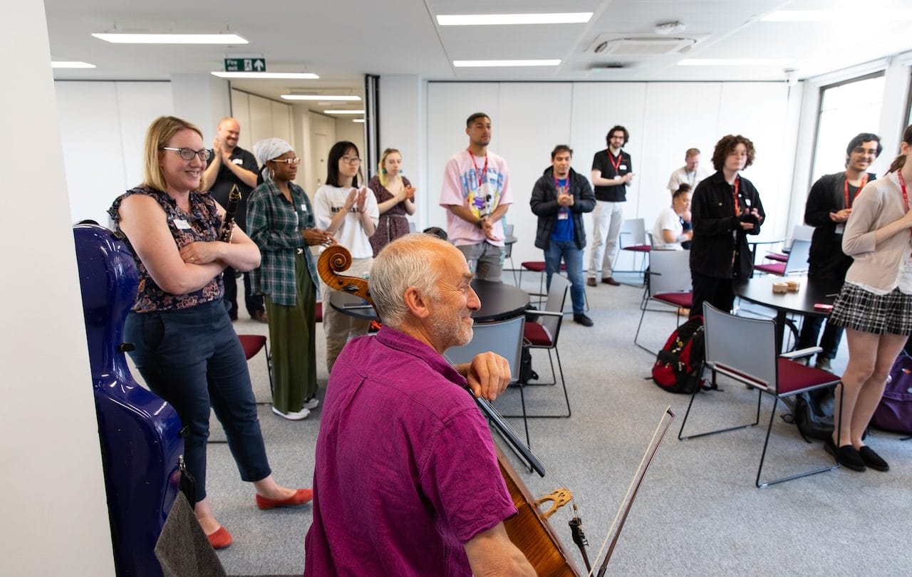 A group of students and a man holding a cello stand in a room together.